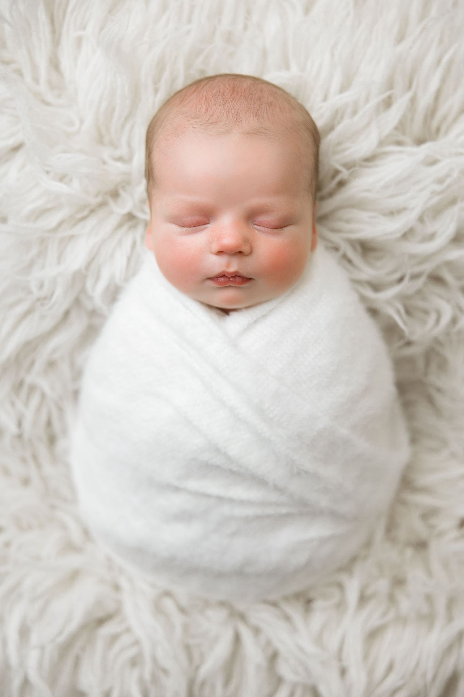 Sleeping newborn baby swaddled in white blanket lying on soft white fur background.