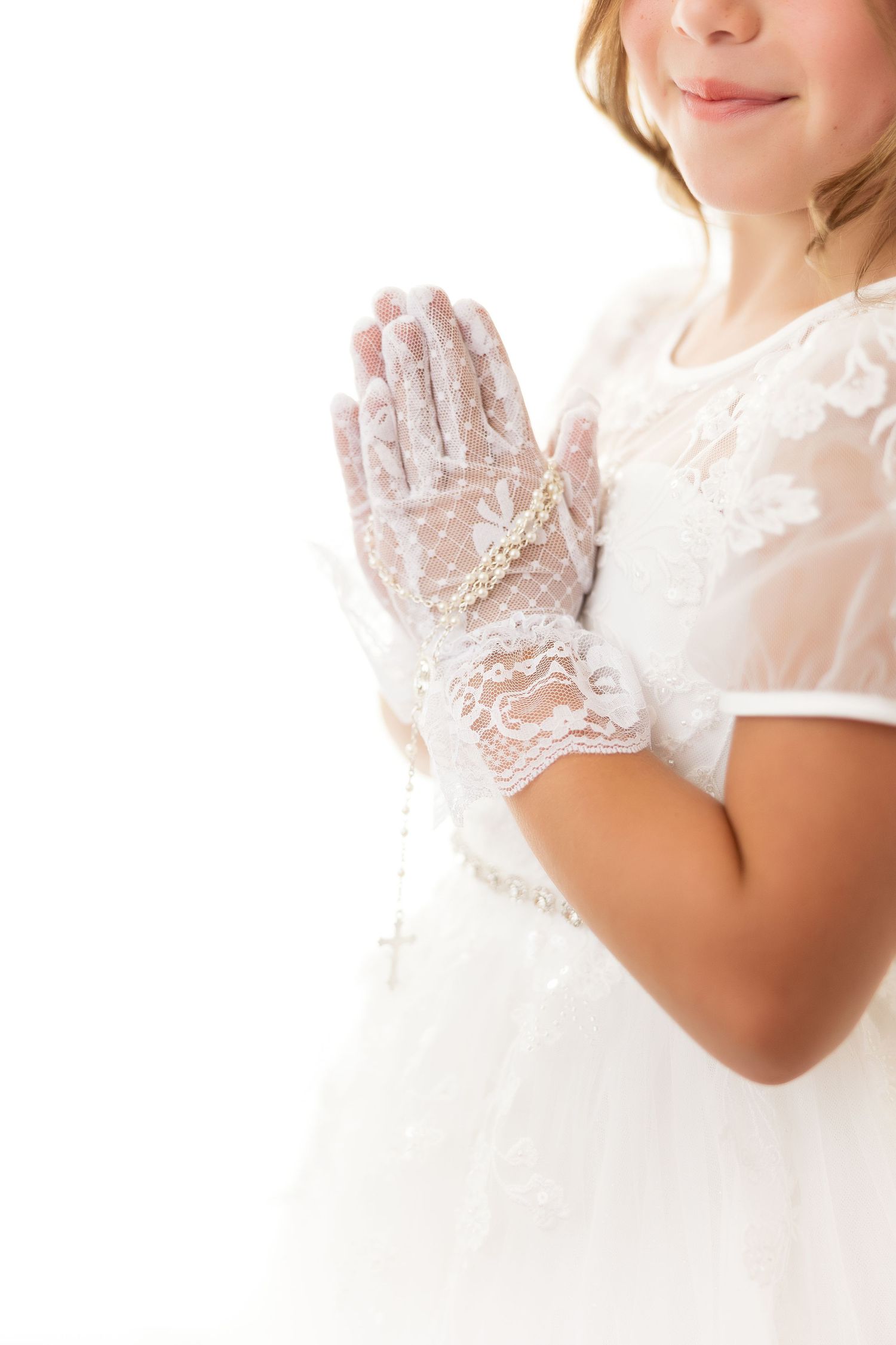 Delicate white lace gloves shown on hands clasped in prayer against a white dress background.
