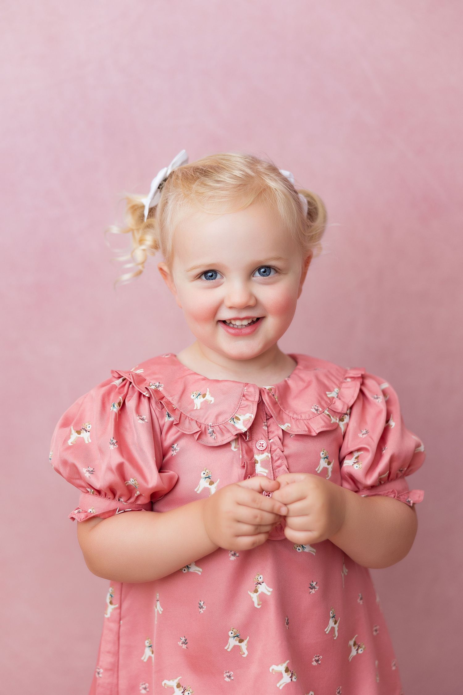 Toddler wearing pink floral dress and white hair bows poses against pink backdrop in portrait studio photo.