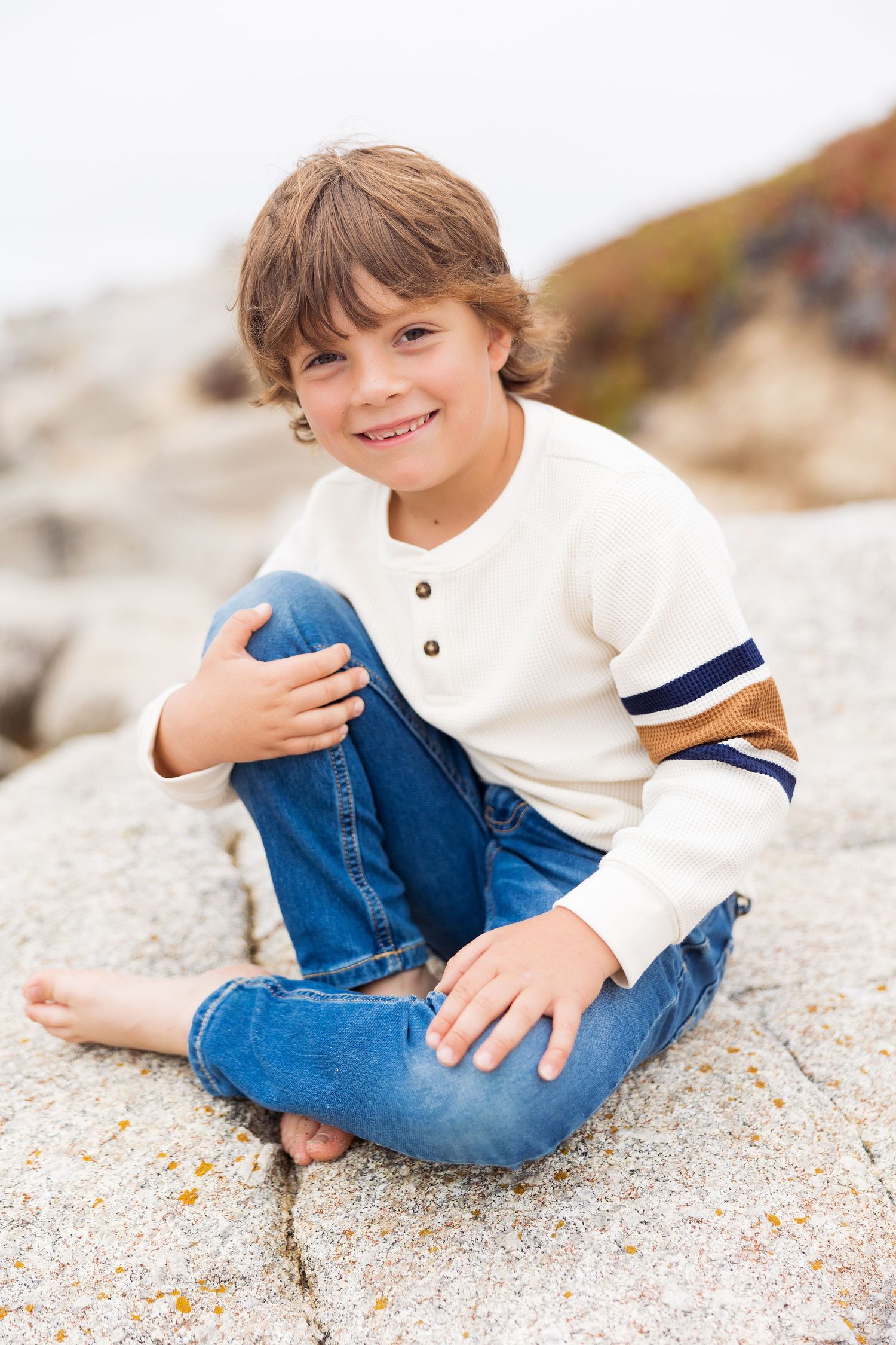 Child in white sweater and jeans sitting casually on sandy beach rocks with natural lighting.