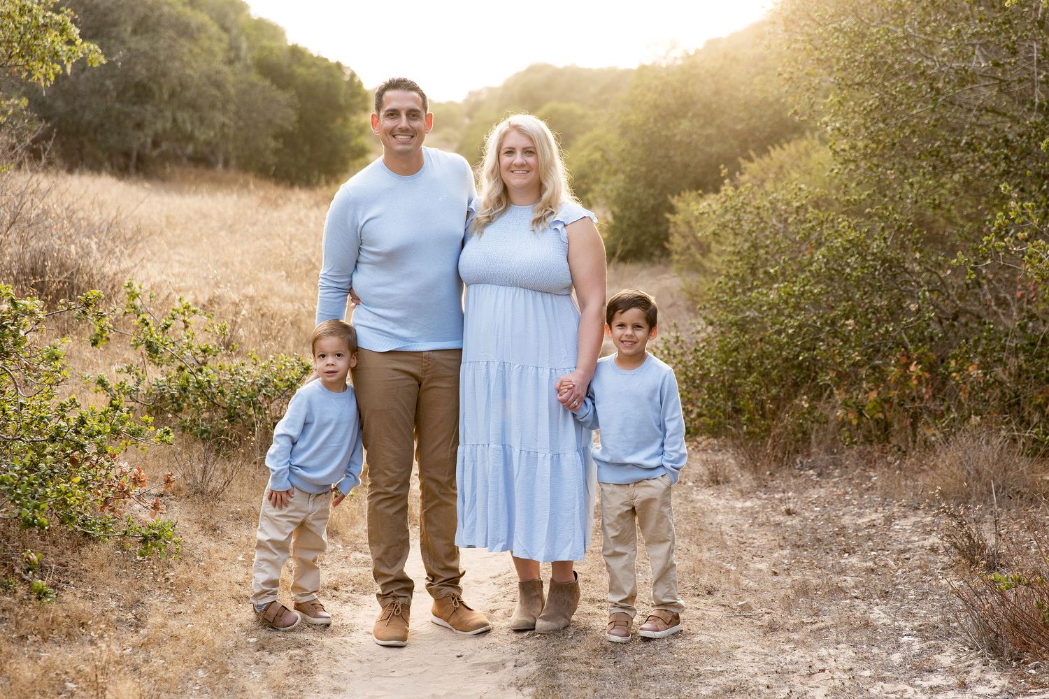 Family portrait on dirt trail wearing coordinated light blue outfits at golden hour.