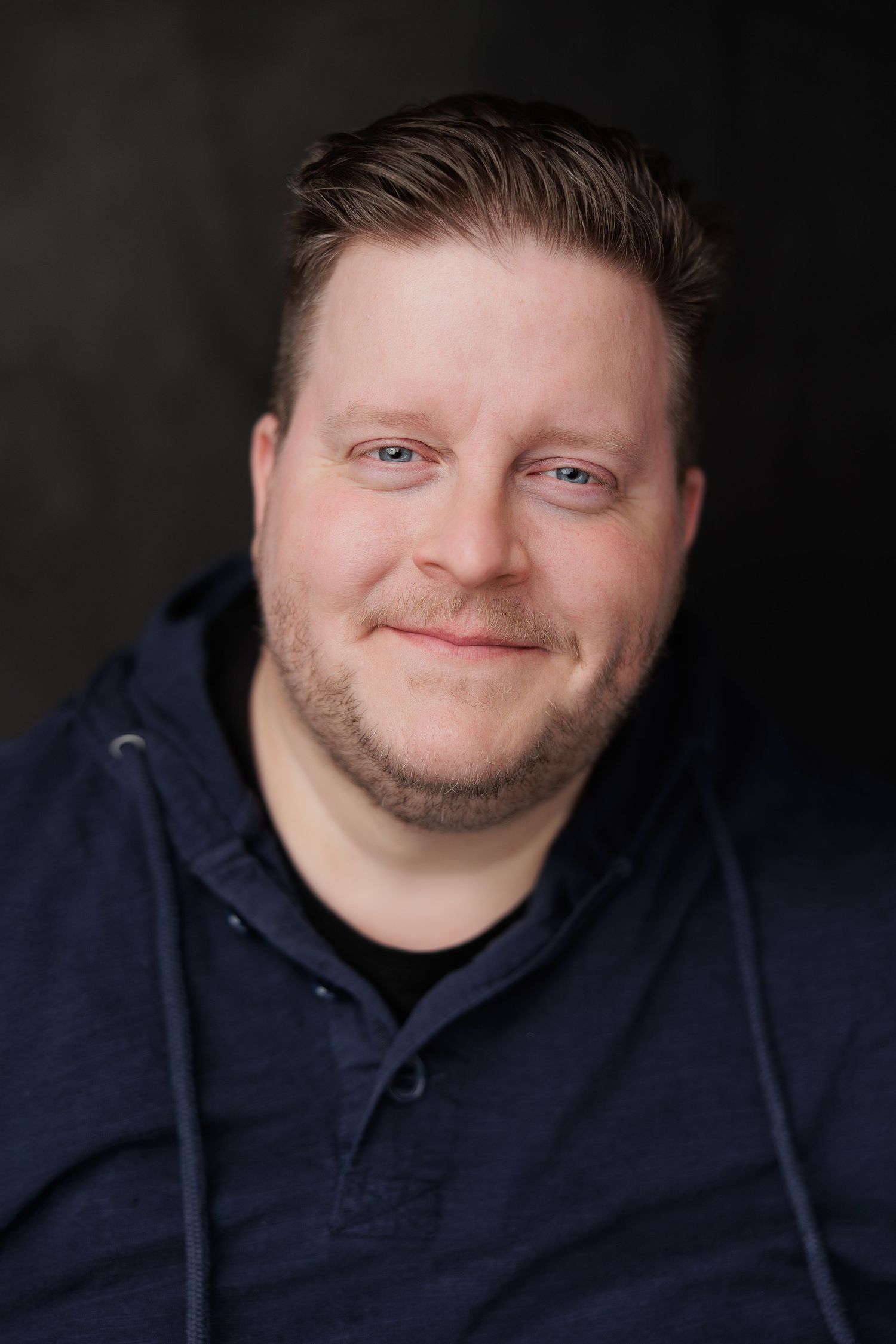 Smiling man with beard wearing a navy blue hoodie against a dark background in a professional headshot photo.