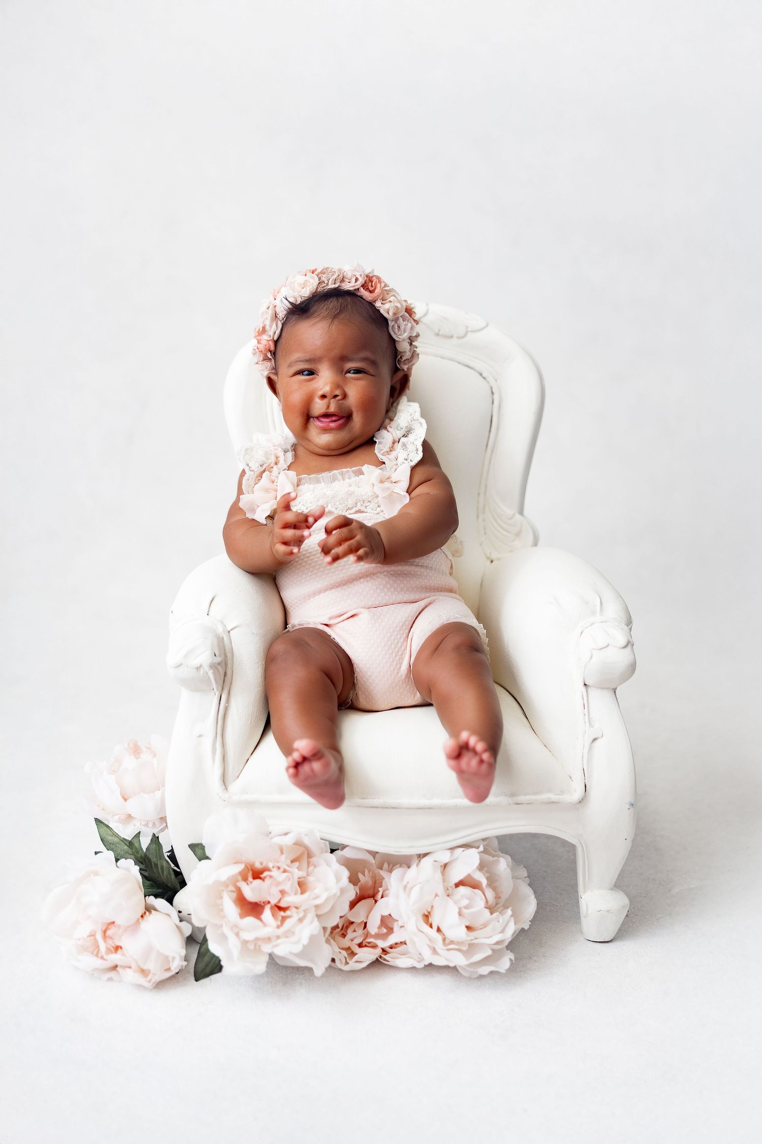 Sweet baby in pink outfit lounges on white ornate chair with delicate peach flowers in studio setting.
