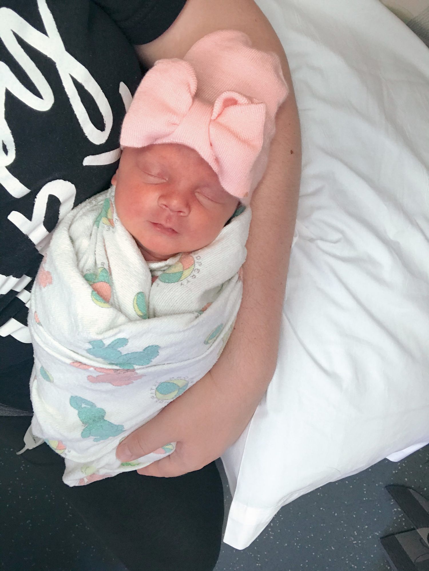 A newborn baby wearing a bow sleeps peacefully while swaddled in white blankets.
