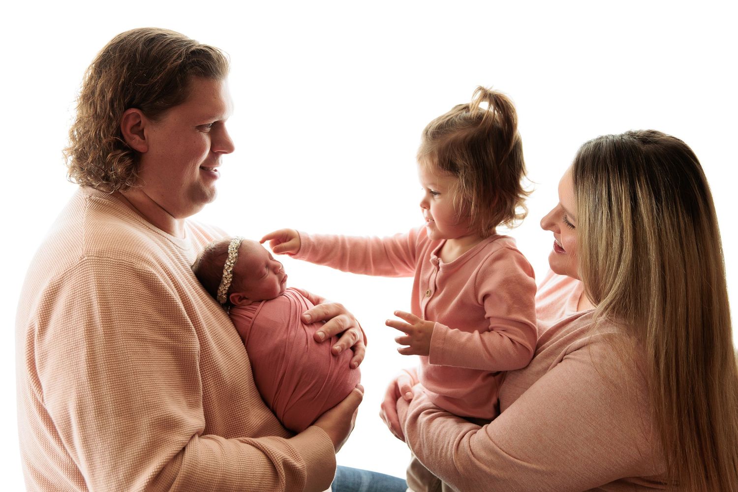 Family in pink and neutral tones sharing tender moment with infant and toddler.