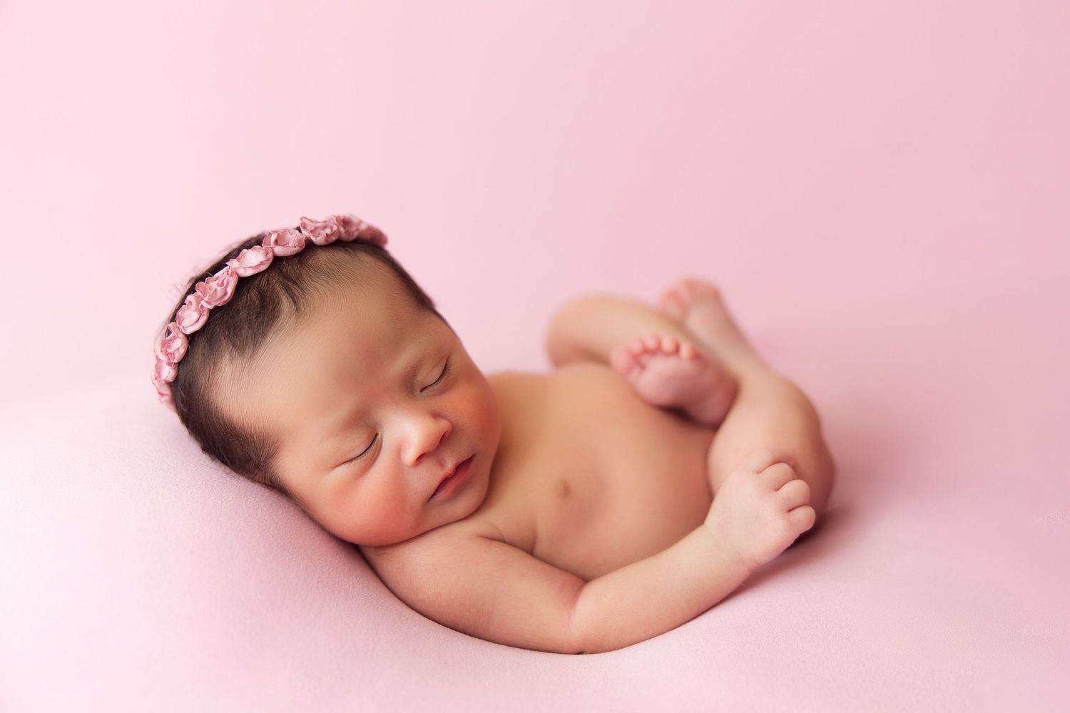 Sleeping newborn with pink flower crown on soft pink background.