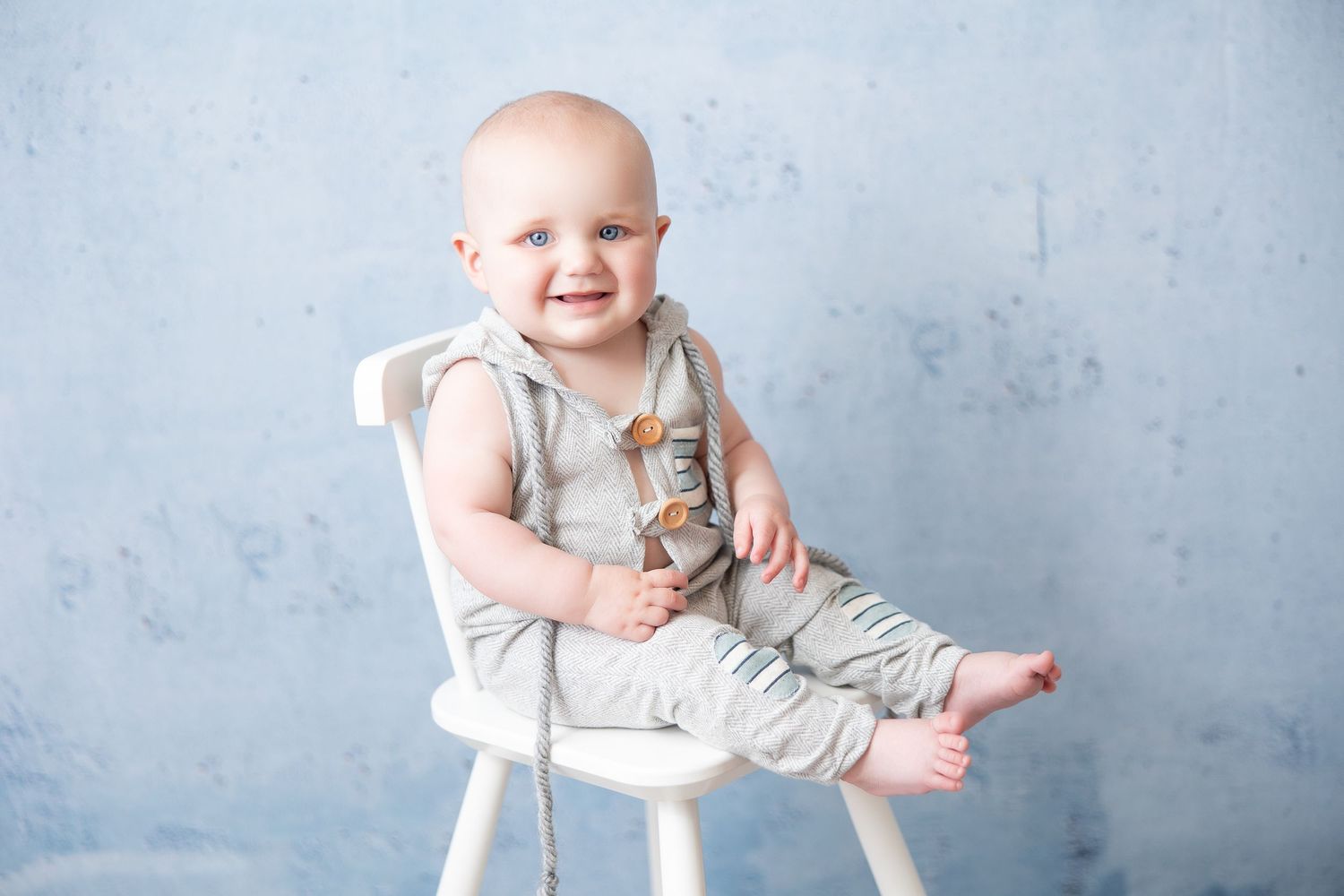 Smiling baby in light overalls sitting on white chair against blue wall.