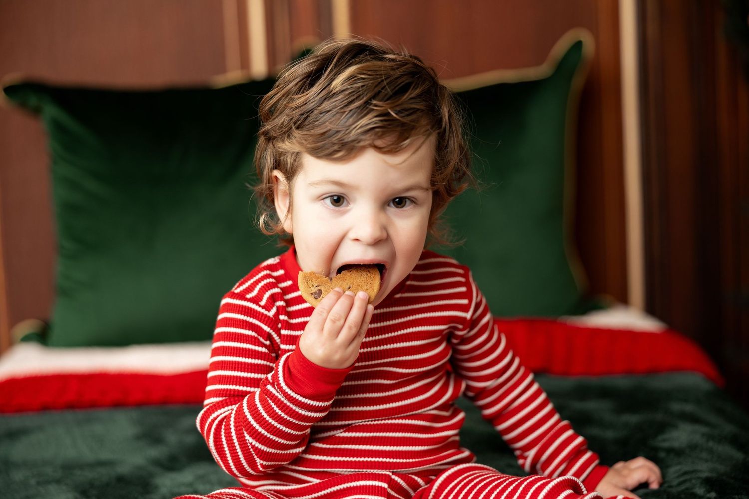 Toddler in striped holiday jammies eating a cookie on a red and green Christmas bed at a Milk & Cookies mini session