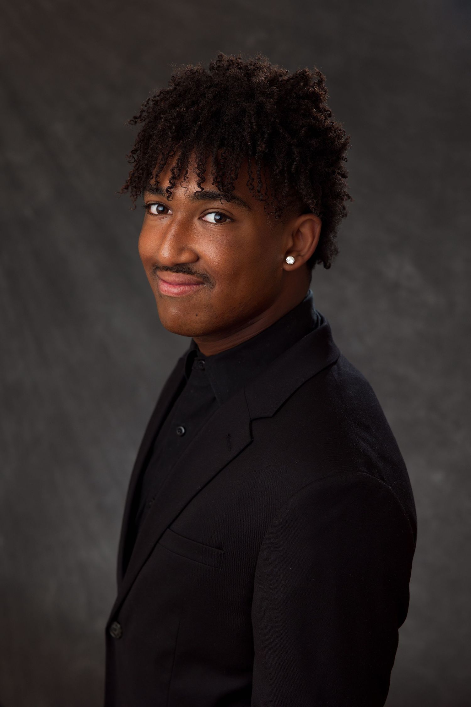 Young man in black suit and turtleneck with curly hair and stud earring smiling in professional portrait.