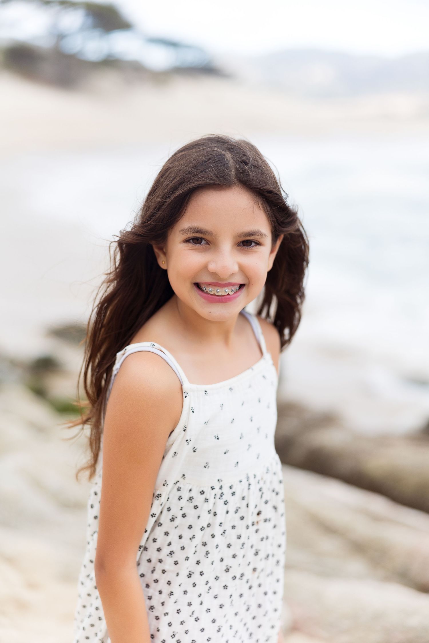 Young girl in white polka dot dress smiles at beach with windswept hair.