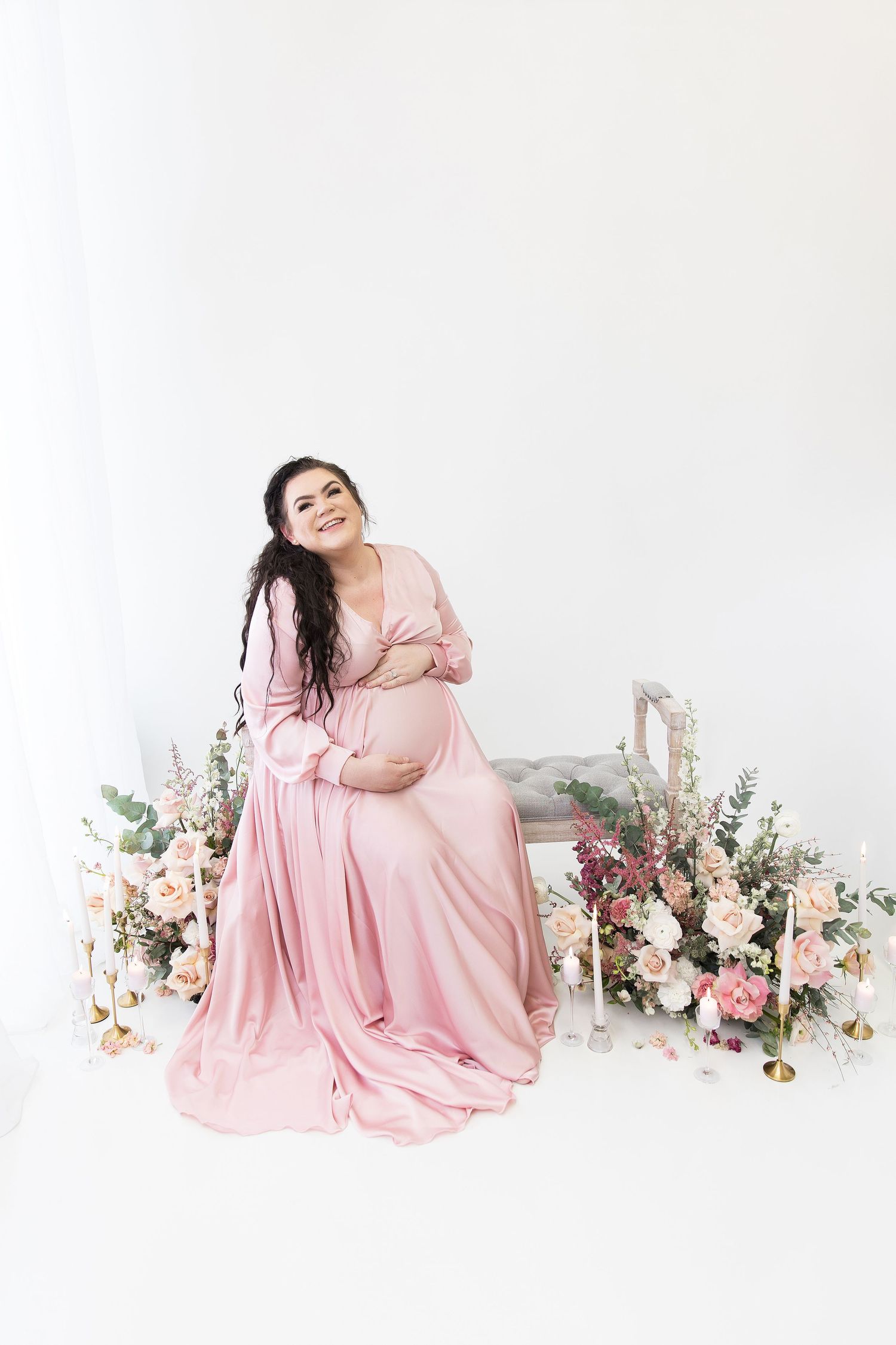 Maternity photos with flowing pink dress and floral decor on white background.