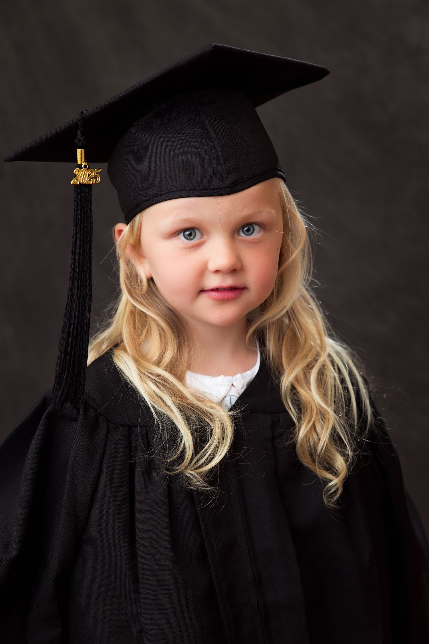 A small child wearing a black graduation cap and gown smiles against a dark background in a series of professional portraits.