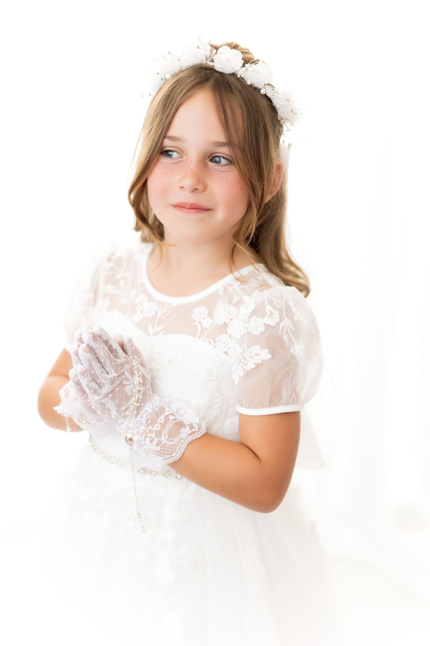 First communion portrait featuring white lace dress, delicate gloves, and floral headpiece against light background.