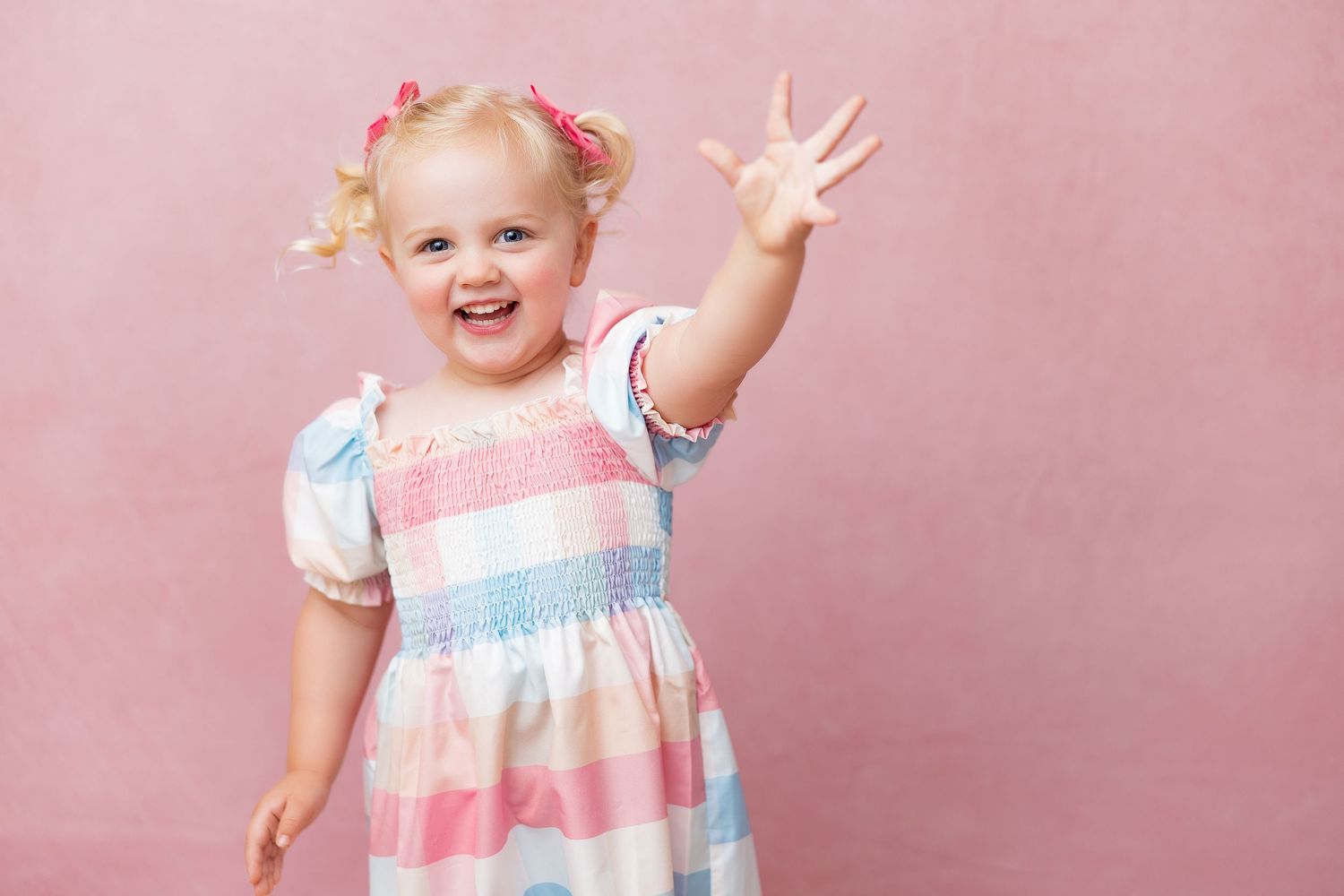 A smiling figure wearing a pink and blue dress poses playfully against a pink backdrop.