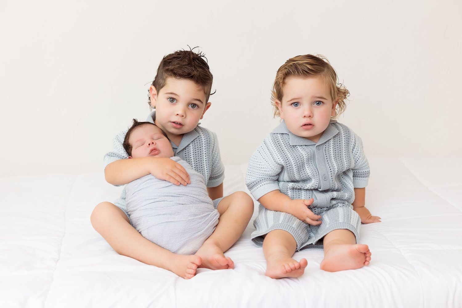 Two children in matching gray shirts sit together on a white bed.