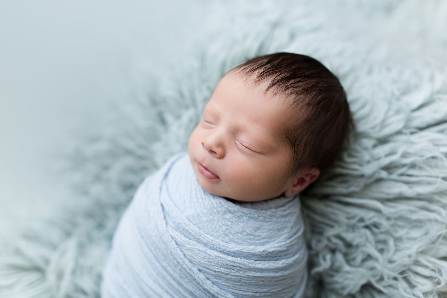 Newborn wrapped in soft grey swaddle on textured light blue backdrop.