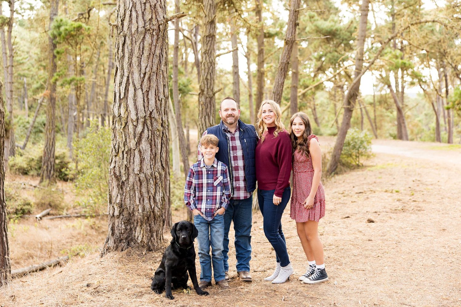 Family of four poses with their black dog on a pine forest trail.