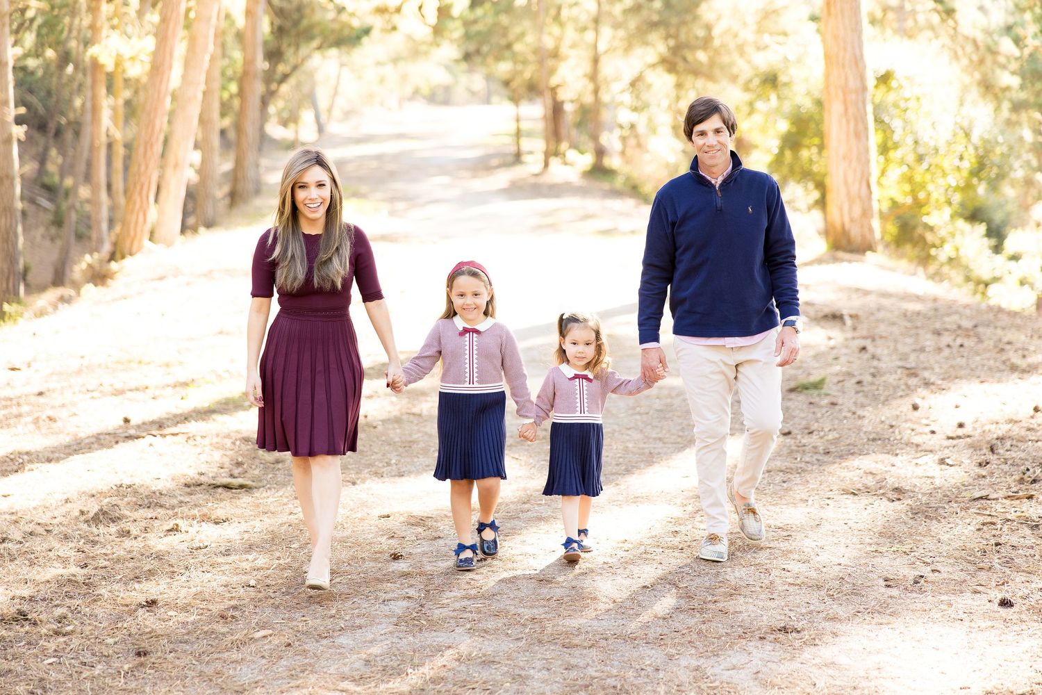 Family walks together down a sunlit wooded trail wearing coordinated navy and burgundy outfits.