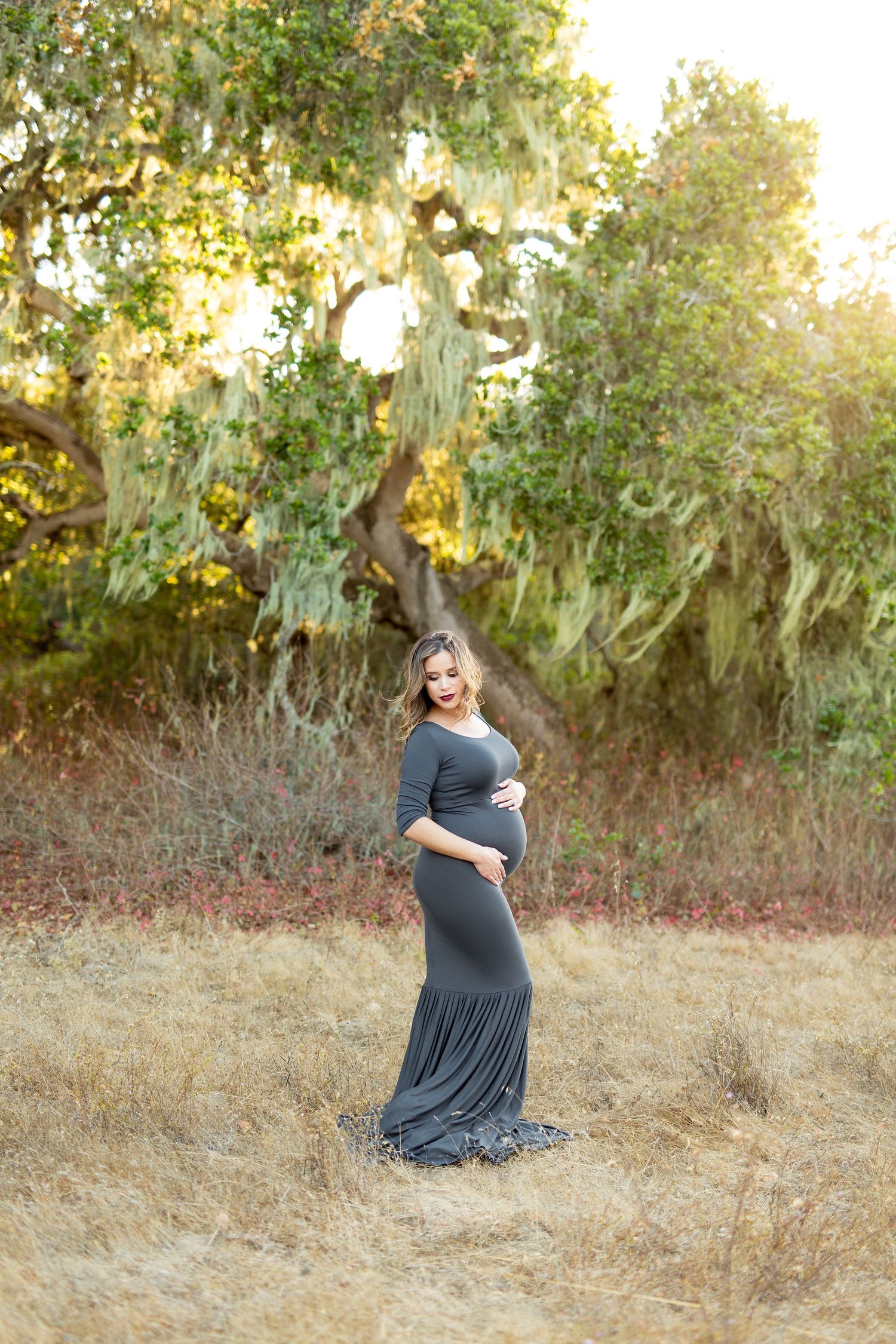 An expectant mother in a flowing navy dress poses gracefully in a golden field at sunset.