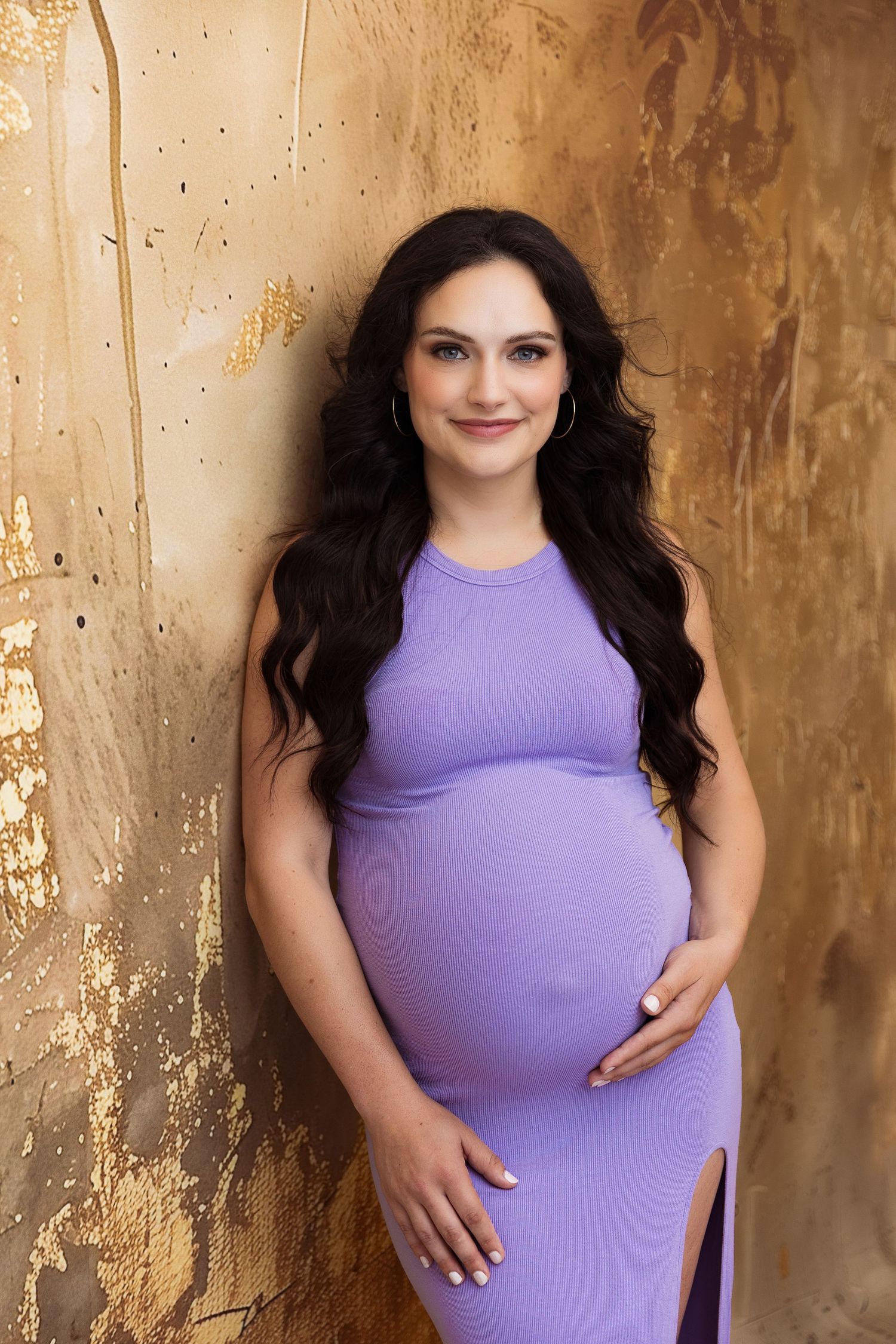 Pregnancy photoshoot with woman in purple dress against textured golden background.