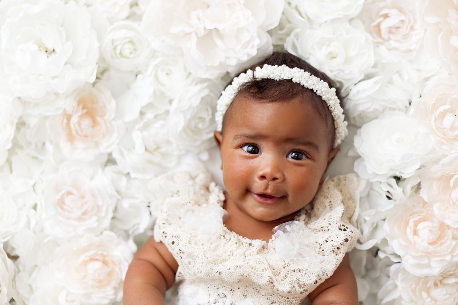 Sweet baby wearing white lace dress and headband lies on soft floral backdrop of pale pink and white roses.