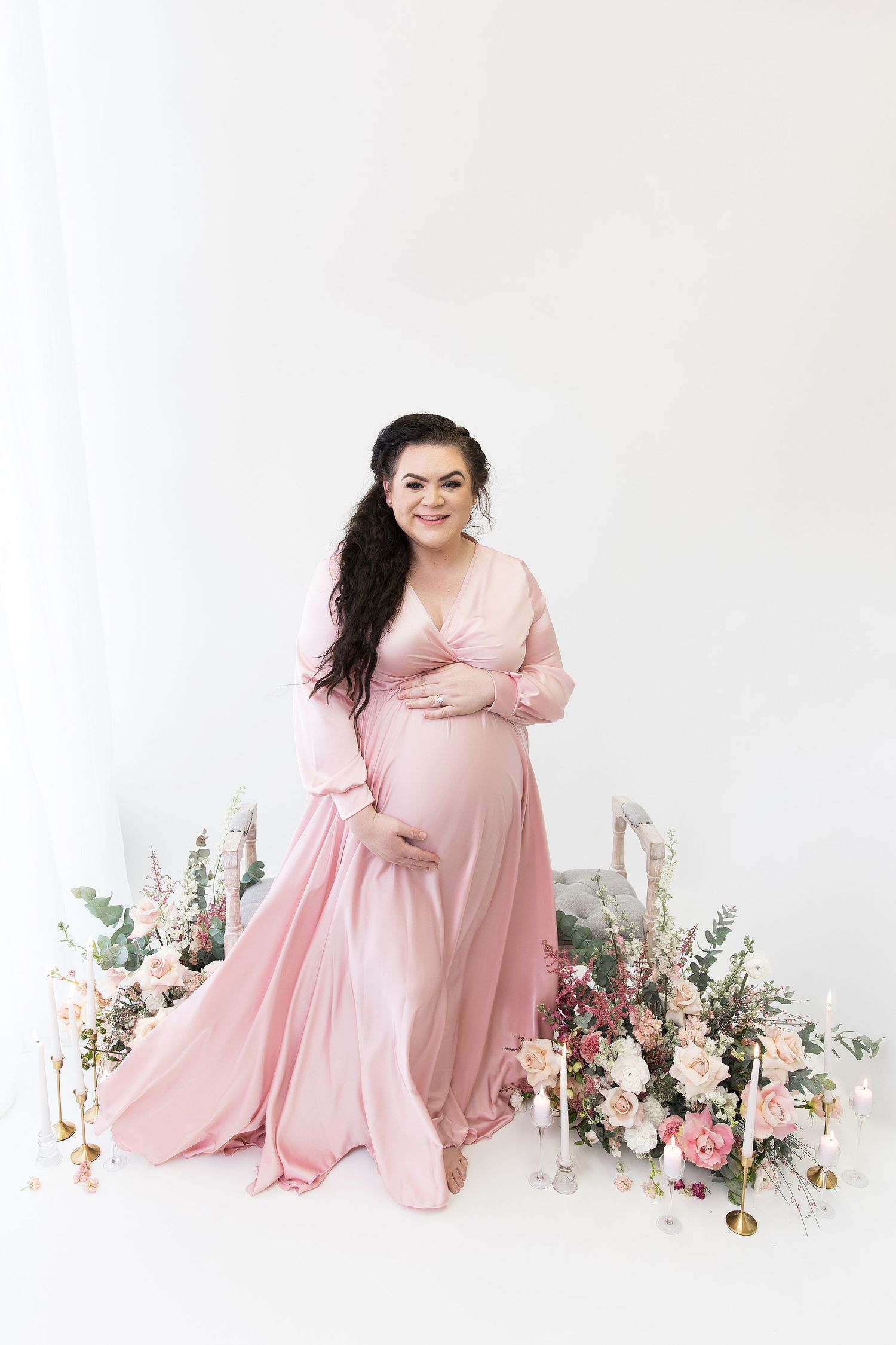 Maternity session with pink dress and floral arrangements on white background.