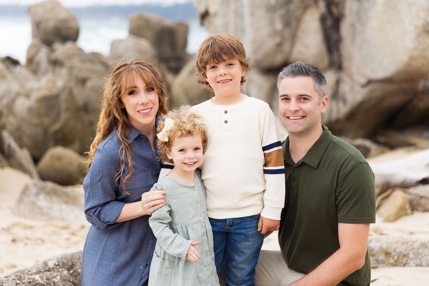 Family in casual clothing poses together on rocky beach during outdoor portrait session.
