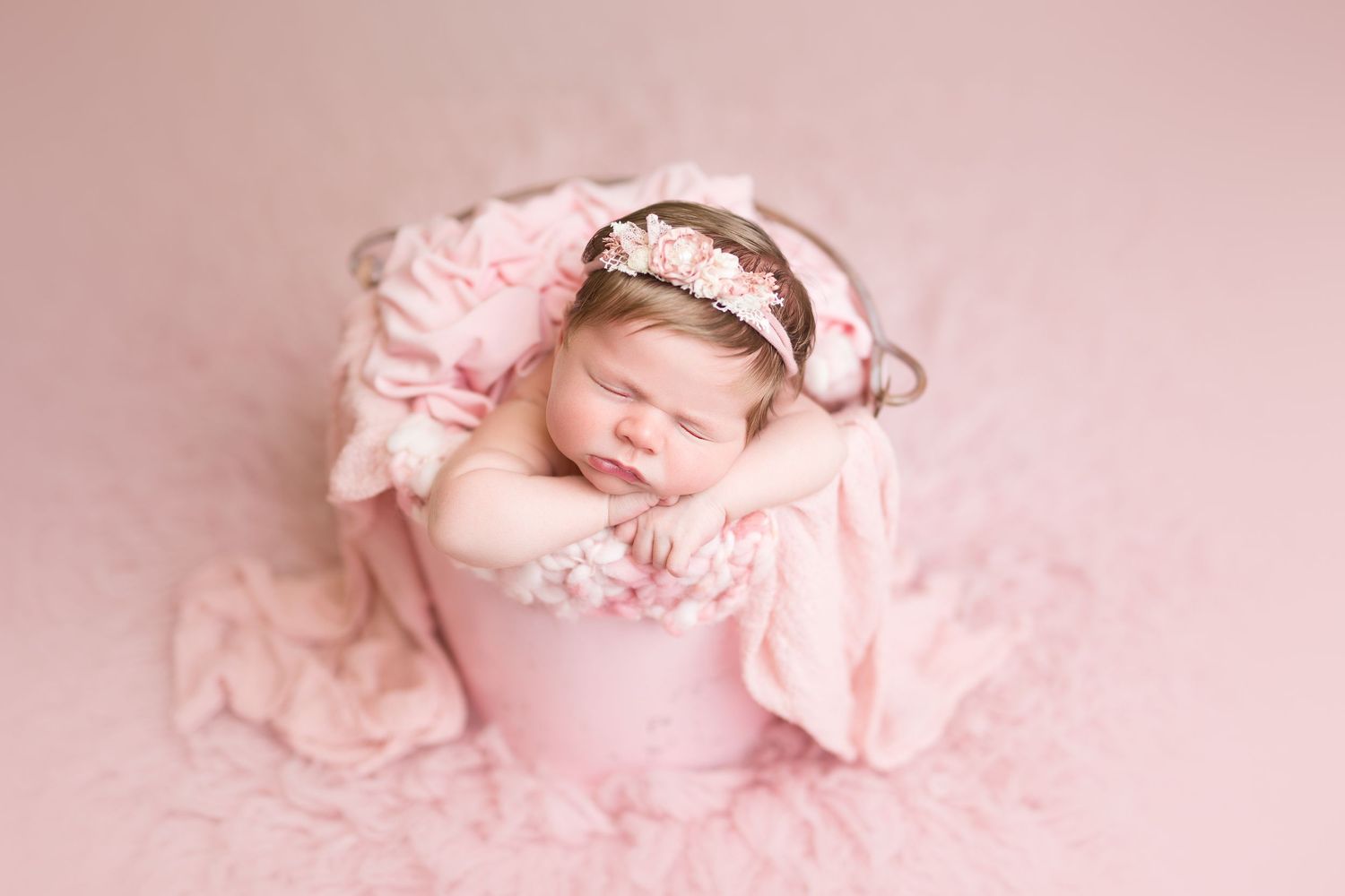 Sleeping newborn in pink headband posed in pink bucket with flowing fabric.