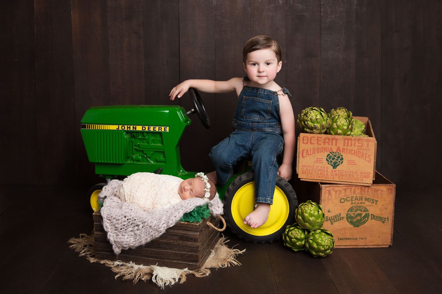 Baby in white romper sleeping near vintage John Deere tractor and wooden produce crates.