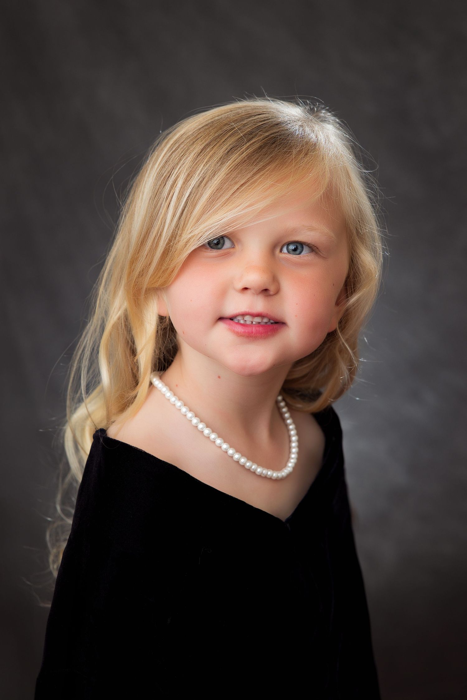 Young child in black dress wearing a pearl necklace poses for elegant portrait against dark background.