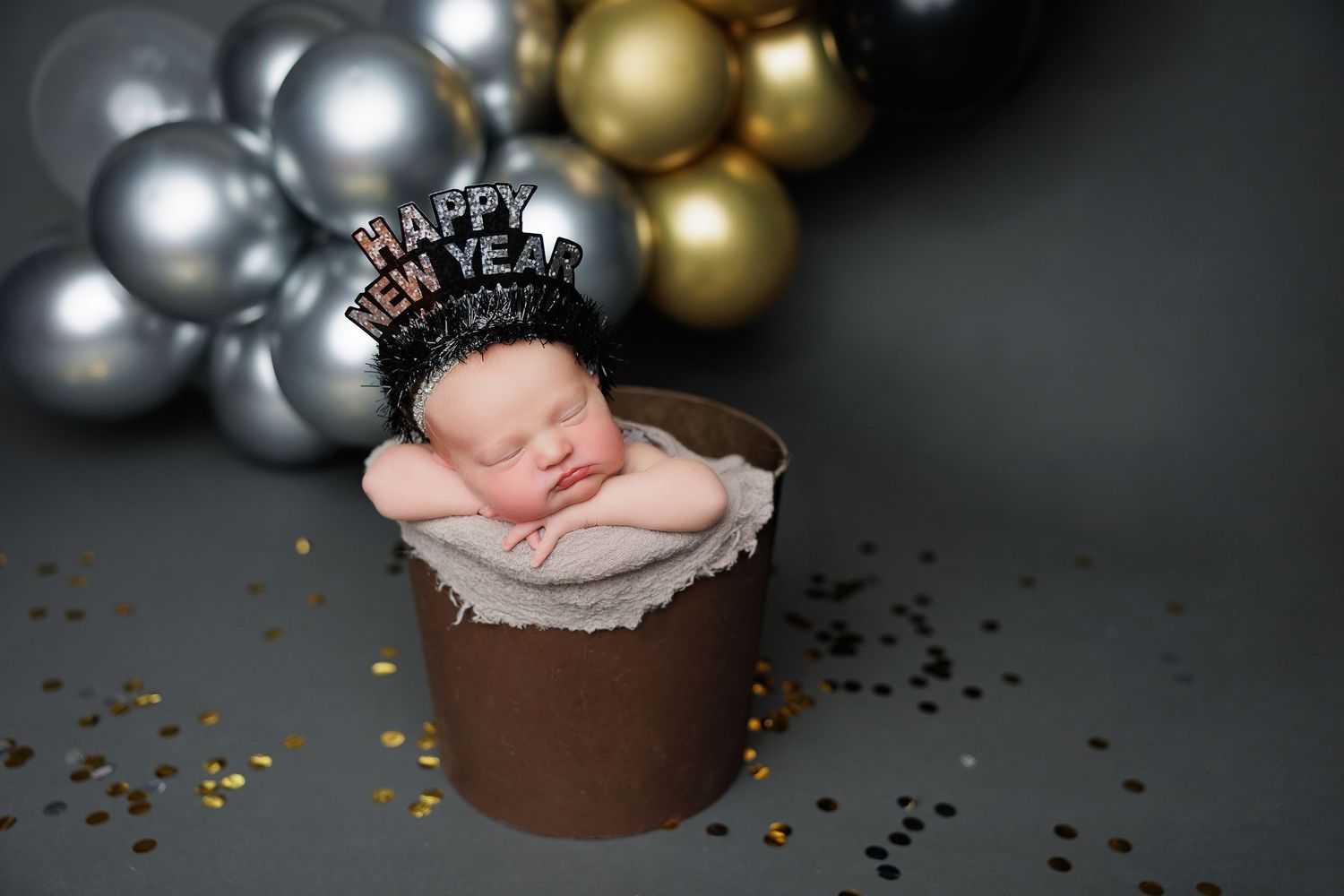 Newborn baby sleeping in a prop bucket wearing a Happy New Year crown, surrounded by gold and silver balloons at Pacific Grove photography.