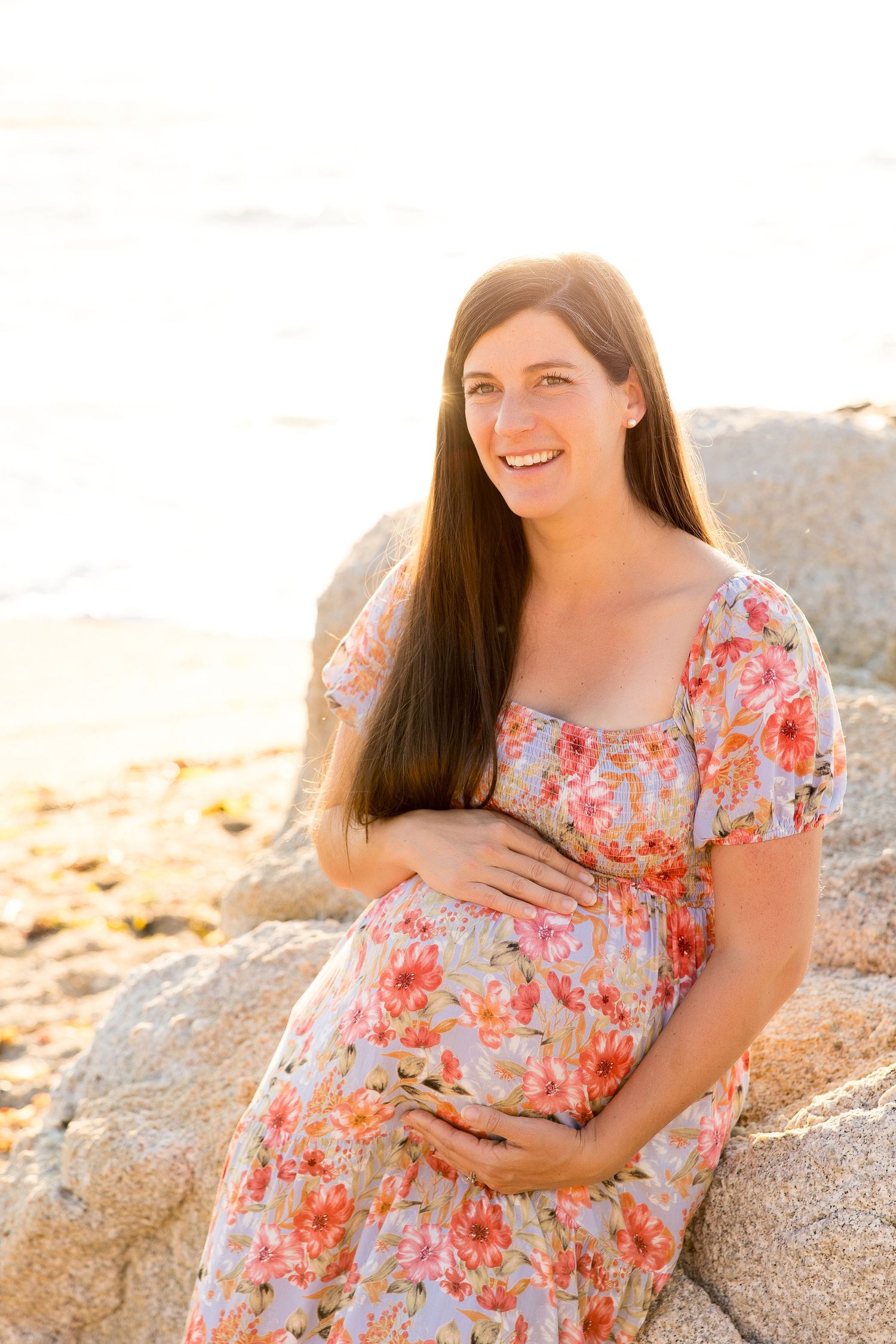 Woman in coral floral dress cradles baby bump during sunset maternity photoshoot.