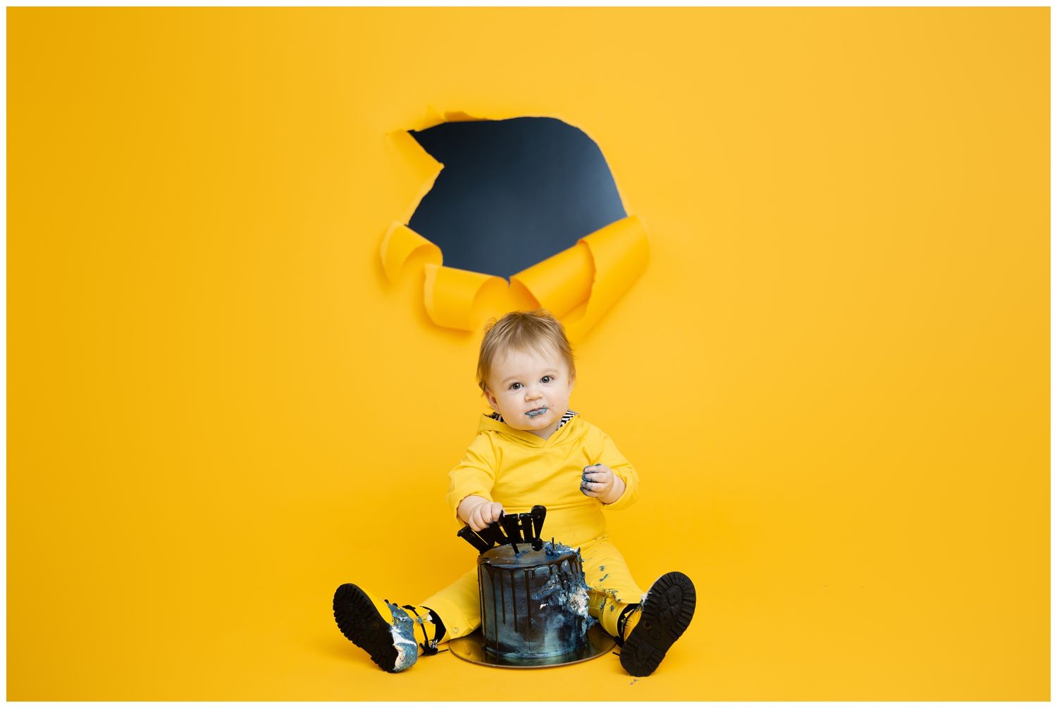 Graduate in blue cap and gown sitting cross-legged against bright yellow background for graduation photos.