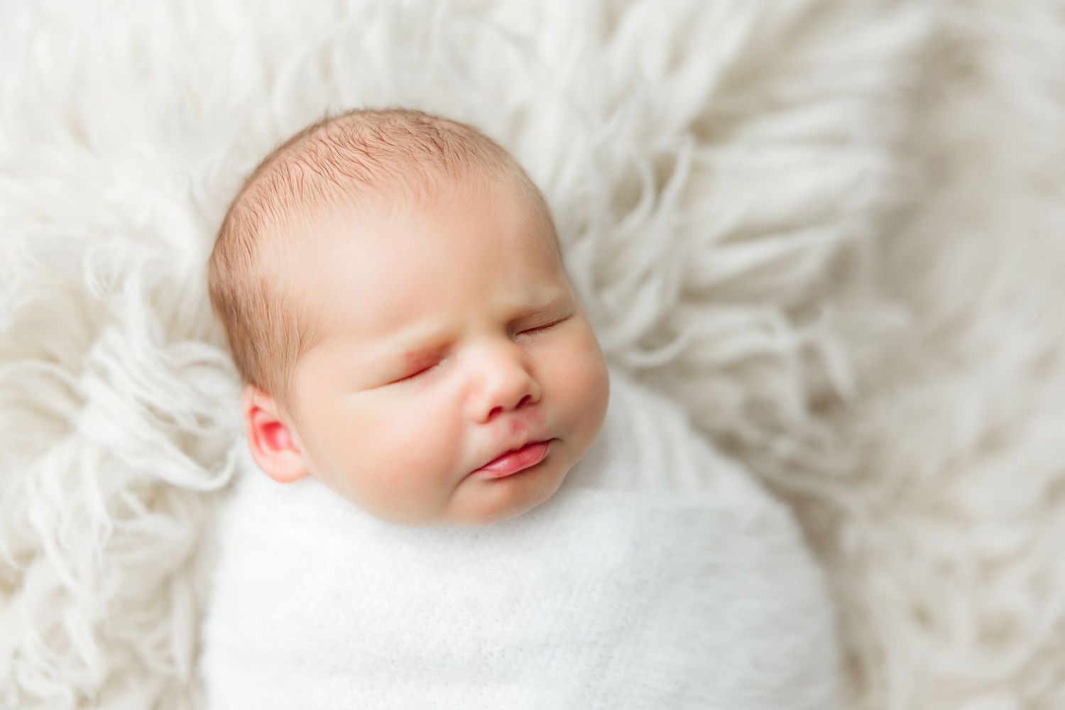 Sleeping baby wrapped in soft white blanket with peaceful expression rests on cozy textured fabric.