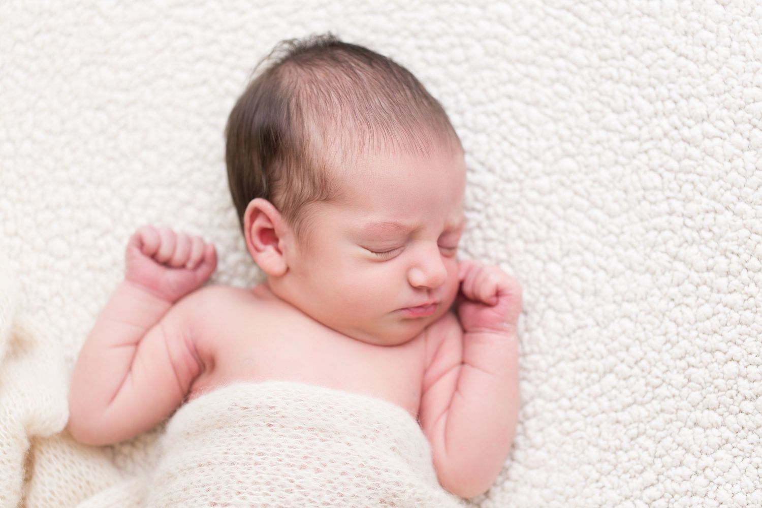Newborn baby sleeping peacefully on white textured surface in muted natural lighting.