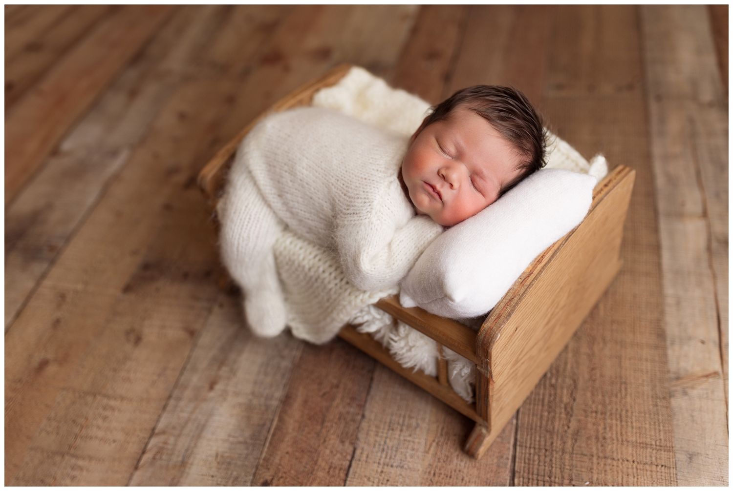 Sleeping newborn wrapped in soft white blanket rests peacefully in wooden bed prop on hardwood floor.