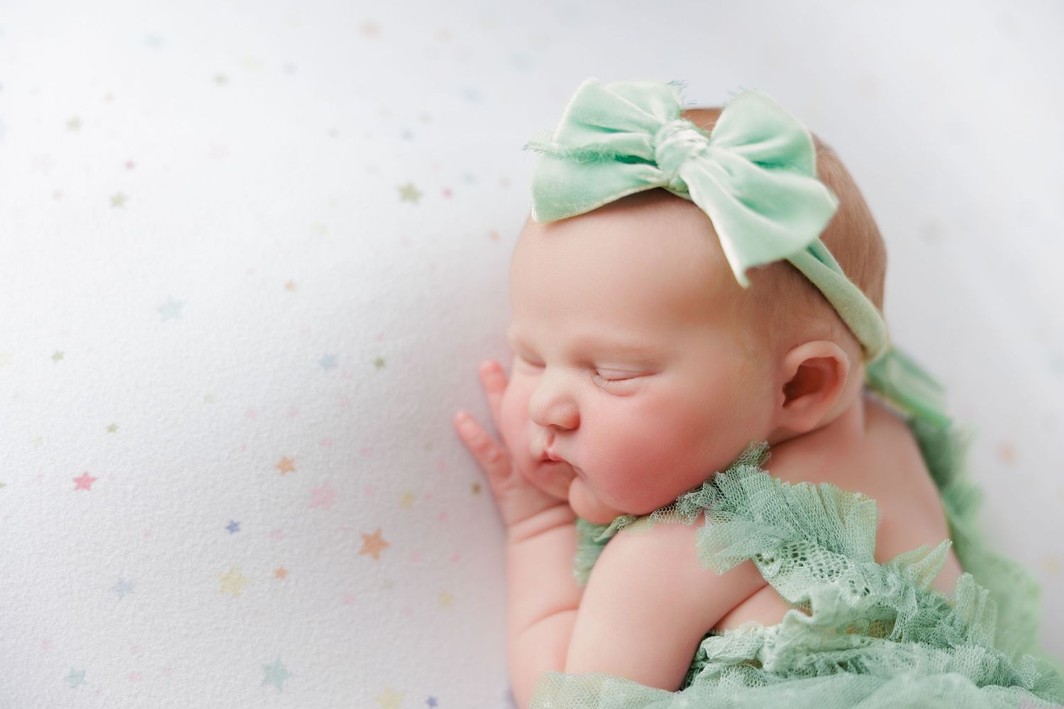 Newborn baby girl sleeping peacefully in a mint green outfit and bow headband on a white star-print background.