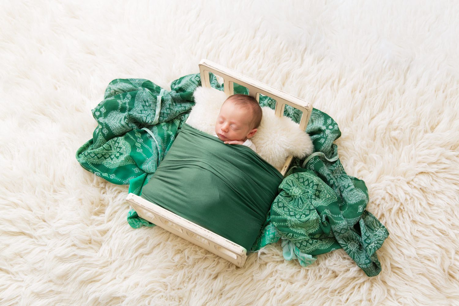Newborn sleeping peacefully on tiny wooden bed with emerald green swaddle and fabric on white fluffy rug.