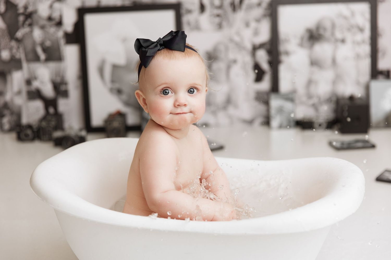 Baby with black bow sitting in vintage white bathtub against wall of photographs.
