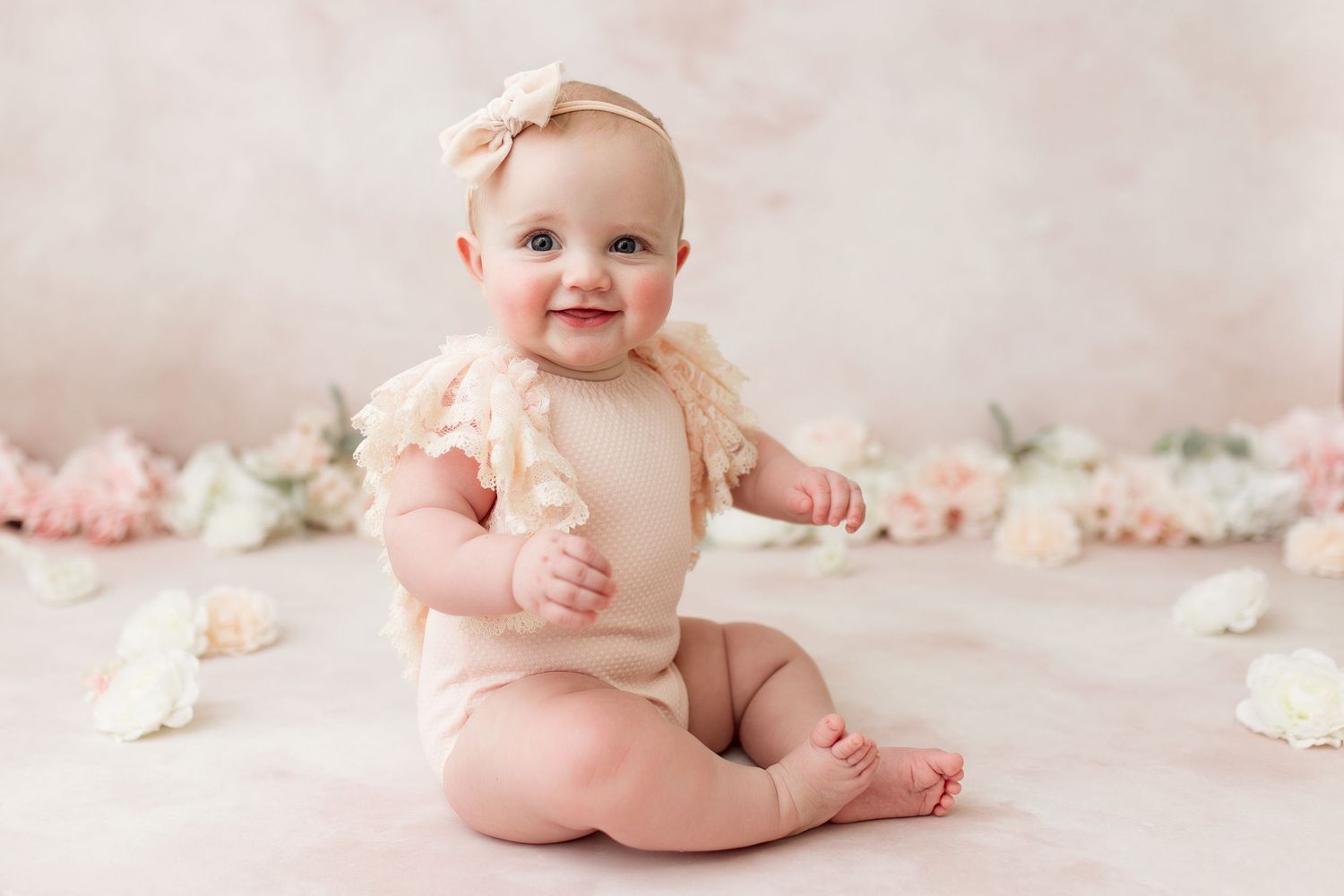 Baby in cream colored romper sitting on pink backdrop with scattered flowers.