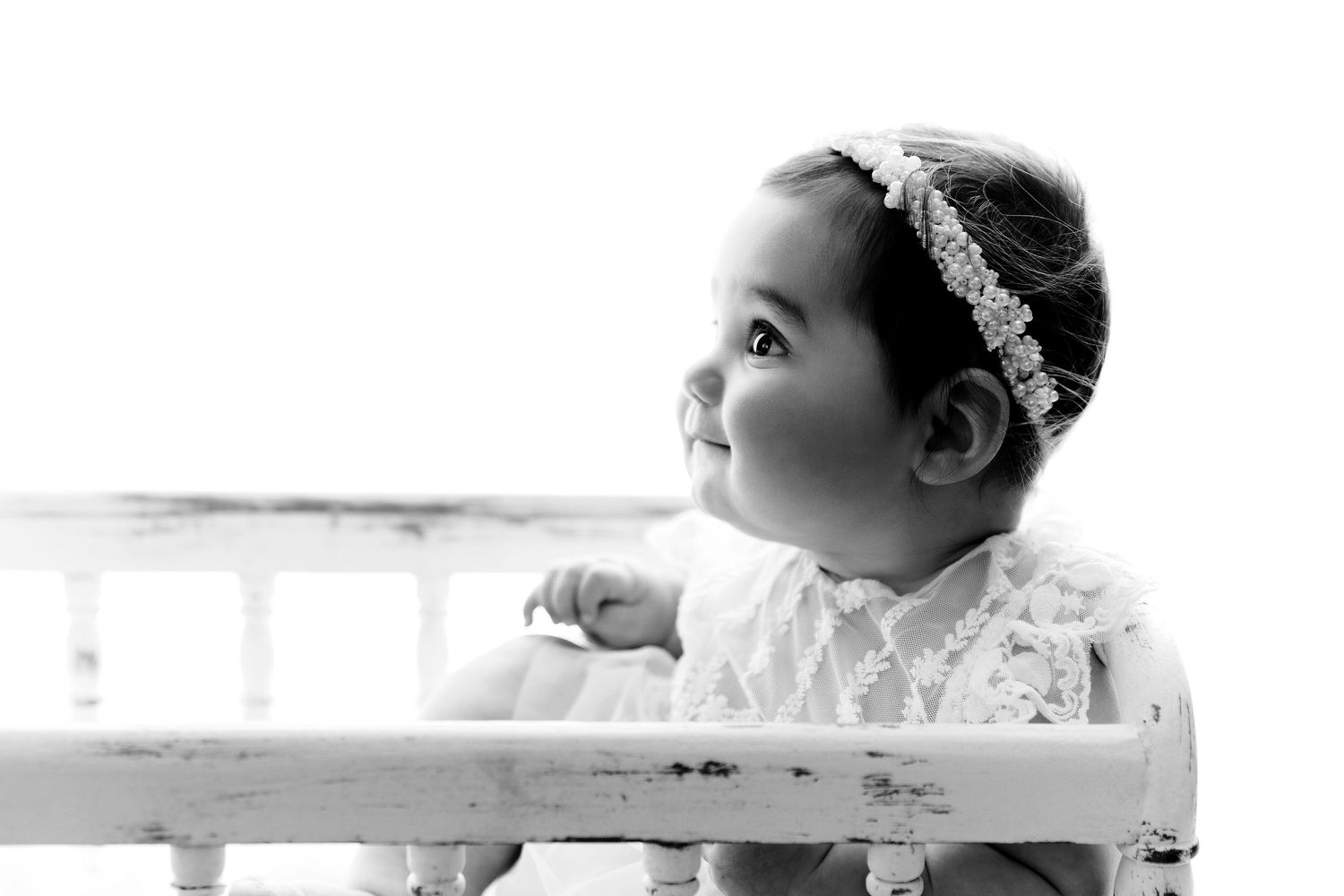 Baby in white lace dress with crystal headband looks up while leaning on white railing.