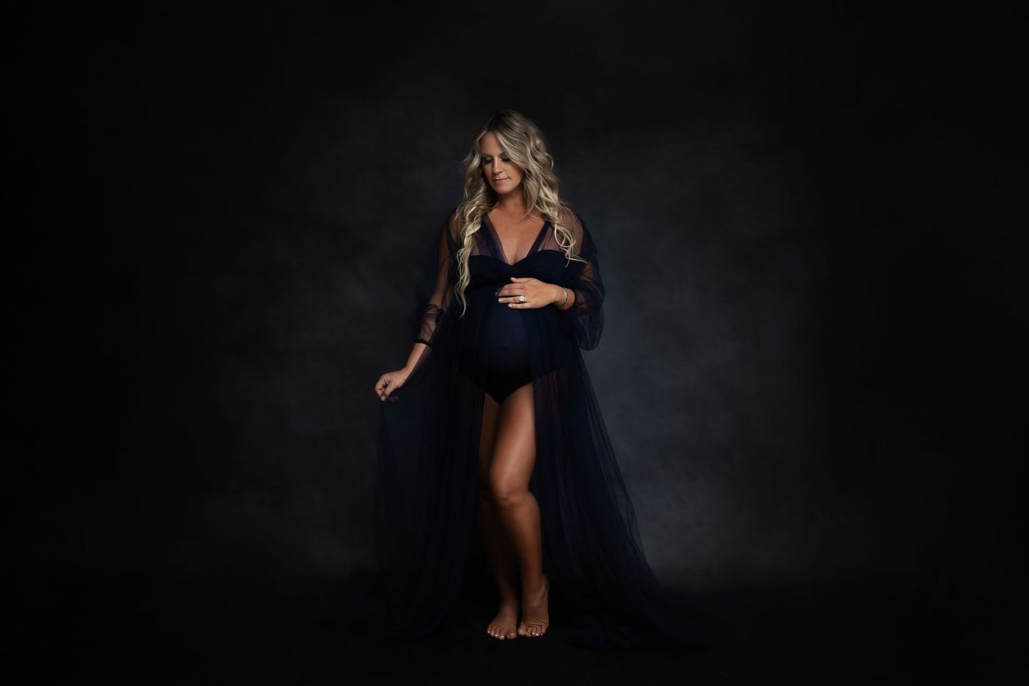 Moody maternity photography session with navy blue gown against dark backdrop.