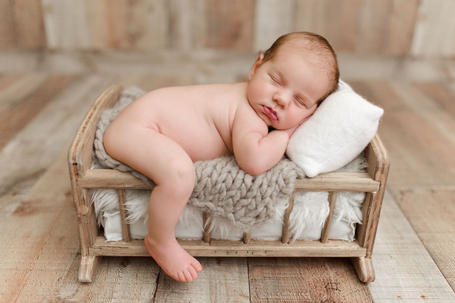Sleeping newborn posed on miniature wooden bed with white pillow.
