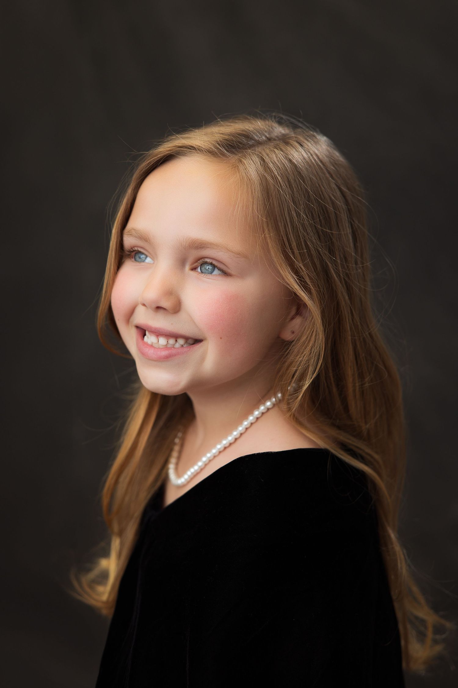 Portrait of a smiling child with long brown hair wearing a black dress and pearl necklace against dark background.