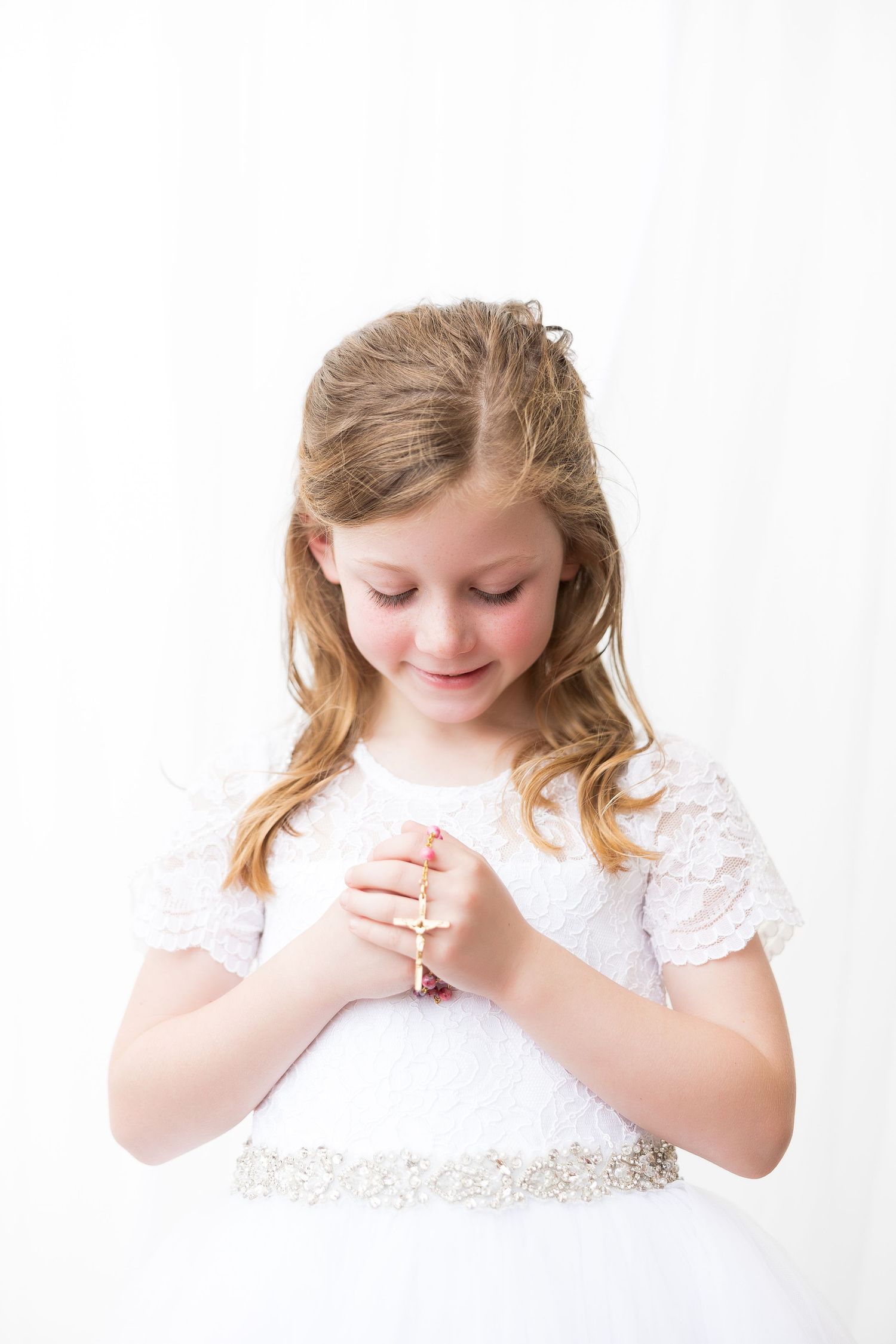 Child in white lace dress looking down at hands with gentle expression.