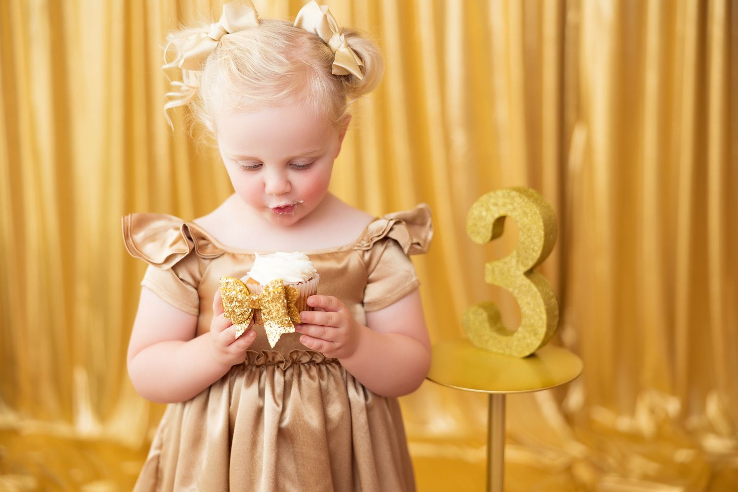 Young child wearing a gold sequin dress stands against a shimmering gold curtain backdrop.