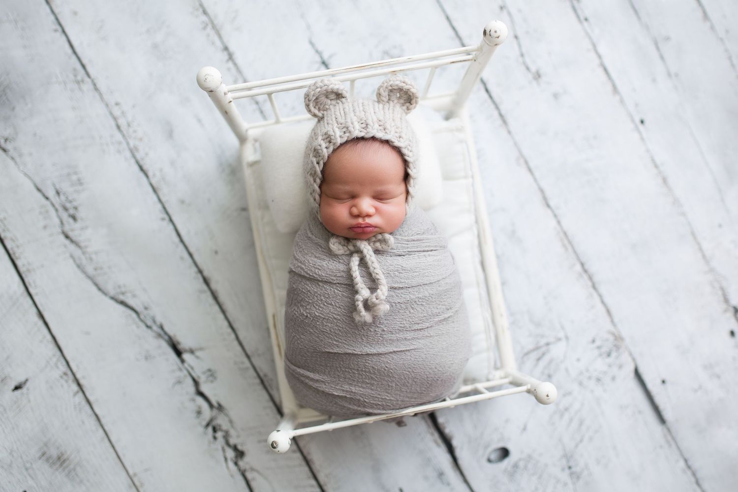 Peaceful newborn sleeping in grey wrap on white wooden bed frame.