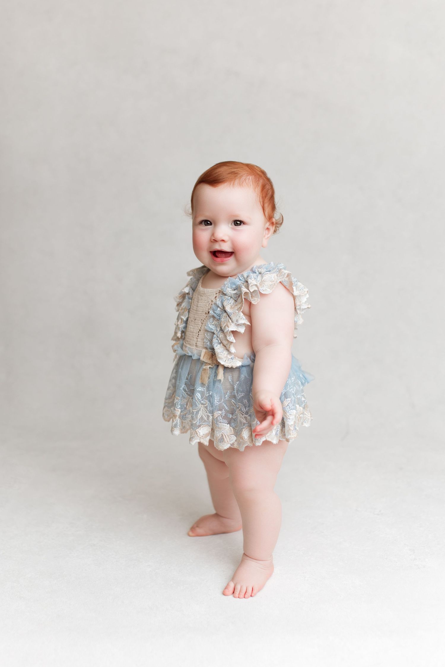 Studio photo of a standing baby wearing a ruffled gray vintage-style romper.