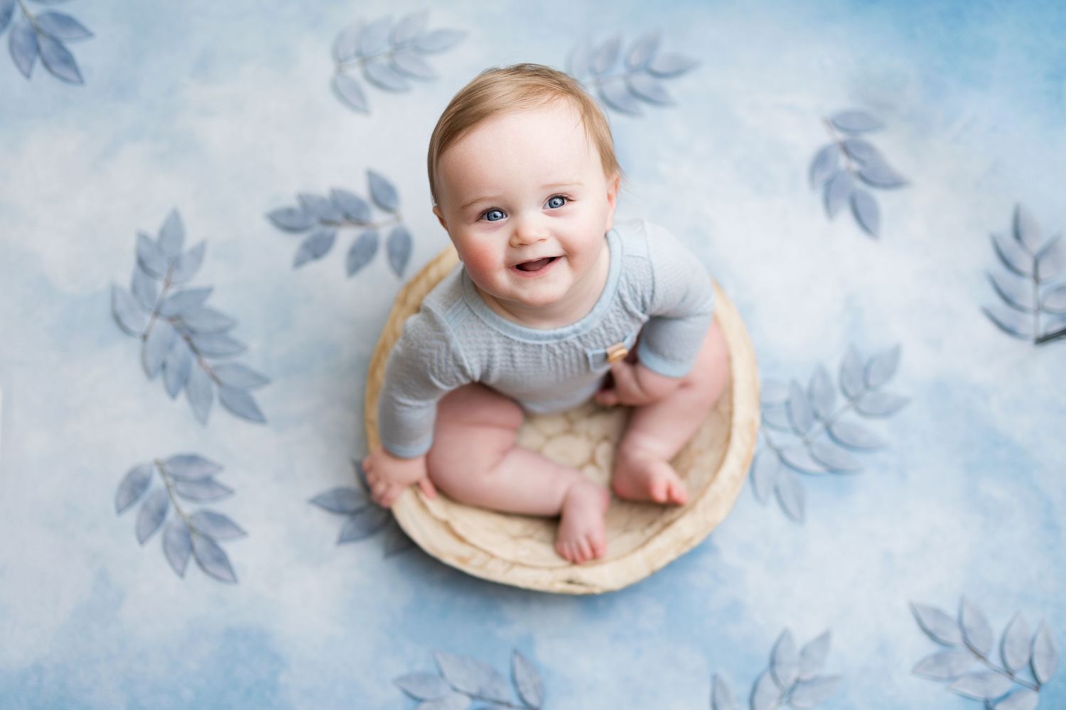 Professional baby photo with blue leaf pattern background and wooden bowl prop.