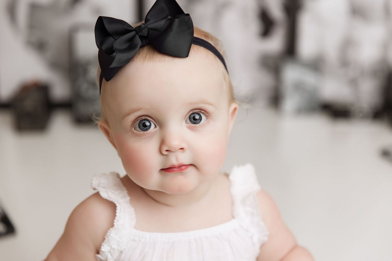 Baby in white dress with black bow posed against photo collage background.
